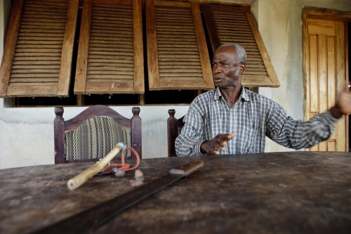 Portrait d'un fermier devant sa maison en Côte d'Ivoire. Sa machette et son lance pierre sont posés sur la table. | Philippe DUREUIL Photographie