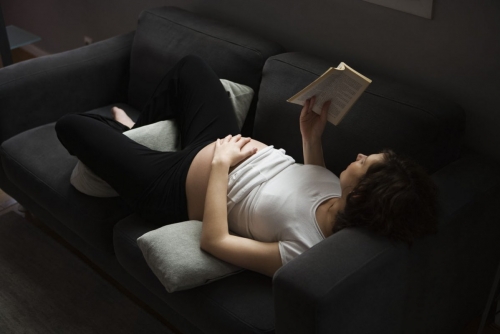 Femme enceinte en train de lire allongée sur un canapé gris vue de dessus | Philippe DUREUIL Photographie